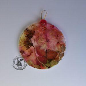 Ręcznie wykonany, okrągły medalion z żywicy, w którym zatopiono różnobarwne suszone rośliny, umieszczony na kartce papieru.