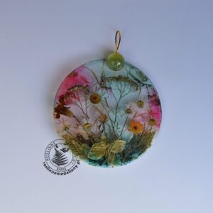 Ręcznie wykonany, okrągły medalion z żywicy, w którym zatopiono różnobarwne suszone rośliny, umieszczony na kartce papieru.